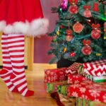 Weihnachtsbaum mit Geschenken. Ein Mädchen in weißroten Socken und einem Weihnachtsrock steht auf den Zehenspitzen.