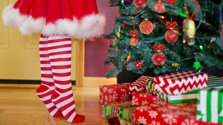 Weihnachtsbaum mit Geschenken. Ein Mädchen in weißroten Socken und einem Weihnachtsrock steht auf den Zehenspitzen.