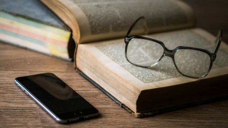 Brille auf einem aufgeschlagenen Buch. Daneben liegt ein Handy.