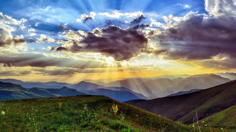 Bild einer Bergwiese auf Bergpanorame. Die Sonne strahlt hinter einer Wolke