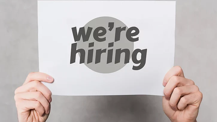 Zwei Hände die ein Schild mit der Aufschrift "We're hiring" in die Luft halten.