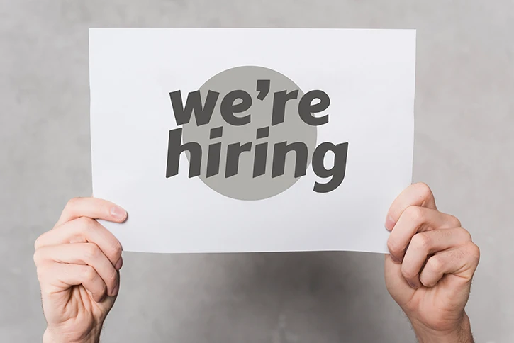 Zwei Hände die ein Schild mit der Aufschrift "We're hiring" in die Luft halten.