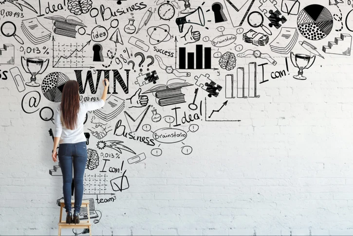 Frau zeichnet divers Symbole, die allesamt mit Statistik, Business, Erfolg, Ideen, etc zu tun haben an die Wand