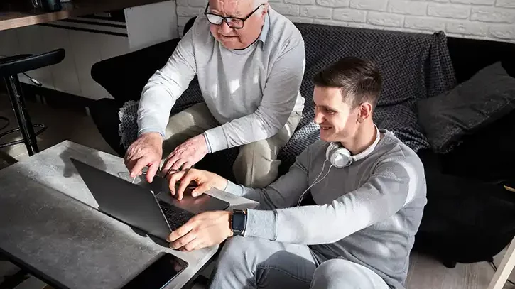 Ein junger Mann sitzt am Boden und erklärt einem älteren Herren, der neben ihm auf der Couch sitzt, etwas an einem Laptop.