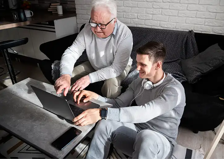 Ein junger Mann sitzt am Boden und erklärt einem älteren Herren, der neben ihm auf der Couch sitzt, etwas an einem Laptop.