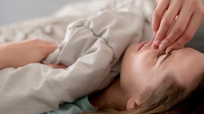 Frau im Bett fasst sich mit schmerzverzerrtem Blick an die Stirn.