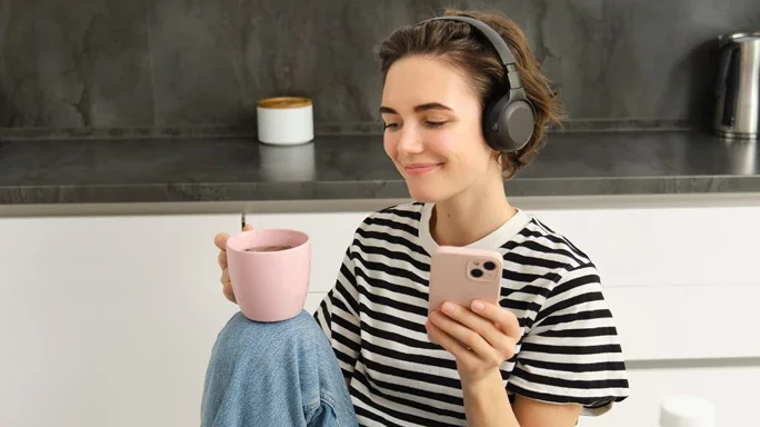 Junge Frau sitzt mit Kaffe und Handy in der Küche und hört sich etwas über Kopfhörer an