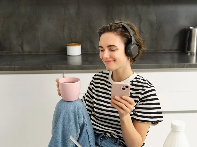 Junge Frau sitzt mit Kaffe und Handy in der Küche und hört sich etwas über Kopfhörer an