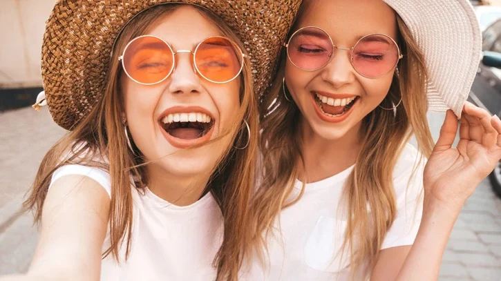 Zwei sommerlich gekleidete Mädchen machen ein Selfie. Sie lachen und haben sehr viel Spaß.