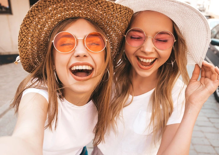 Zwei sommerlich gekleidete Mädchen machen ein Selfie. Sie lachen und haben sehr viel Spaß.