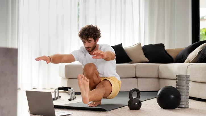 Mann macht zu Hause Fitnessübungen. Vor ihm steht ein Laptop und um ihn herum diverses Fitnesszubehör