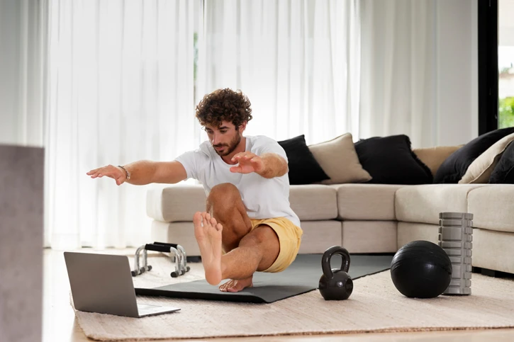Mann macht zu Hause Fitnessübungen. Vor ihm steht ein Laptop und um ihn herum diverses Fitnesszubehör
