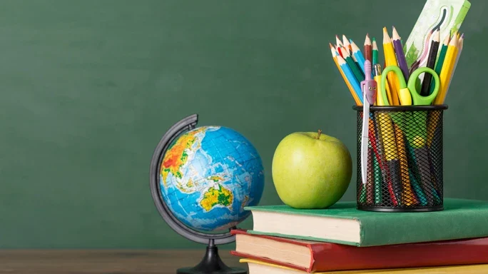 Auf einem Schreibtisch steht ein kleiner Atlas, drei Bücher ein Apfel und ein Becher mit Buntstiften