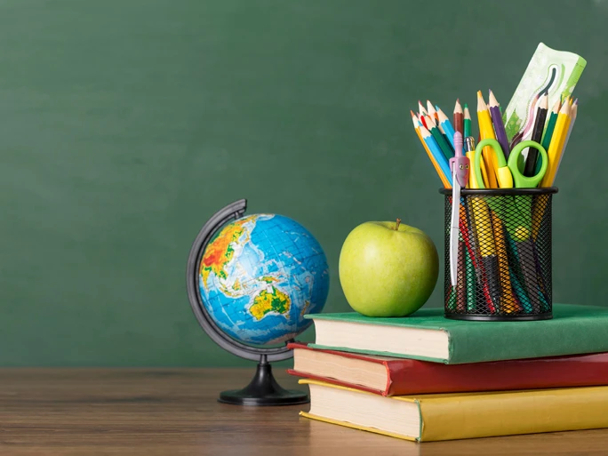Auf einem Schreibtisch steht ein kleiner Atlas, drei Bücher ein Apfel und ein Becher mit Buntstiften