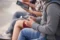 Nahaufnahme von Personen, die in einer Reihe sitzen und diversen mobile Geräten wie Tablets und Smartphones in ihren Händen halten und bedienen.