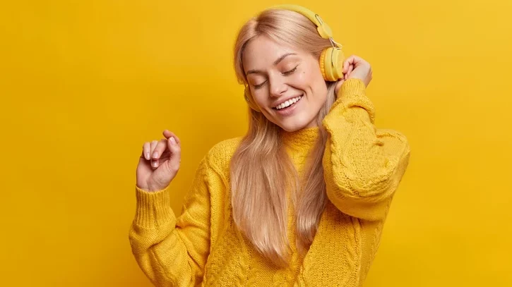 Blonde Frau mit einem gelben Pulli und gelben Kopfhörern tanzt zur Musik vor einem gelben Hintergrund