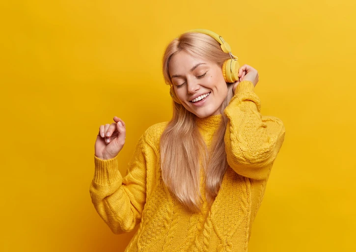 Blonde Frau mit einem gelben Pulli und gelben Kopfhörern tanzt zur Musik vor einem gelben Hintergrund