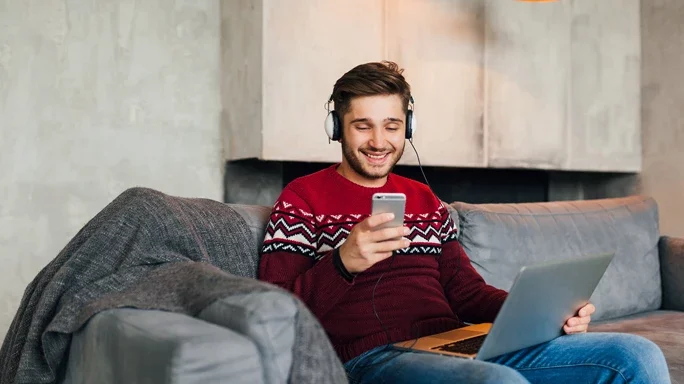 Mann sitzt auf Couch mit Laptop und Mobiltelefon. Er lacht.