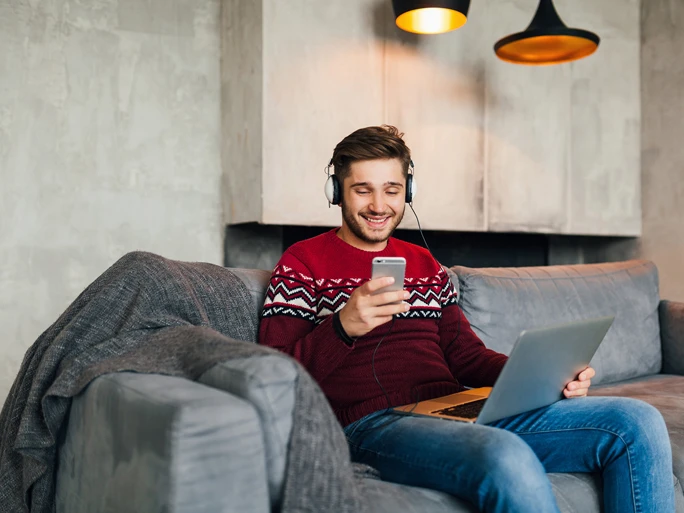 Mann sitzt auf Couch mit Laptop und Mobiltelefon. Er lacht.