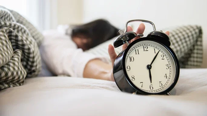Symbolbild zum Beitrag Probleme nach der Zeitumstellung - Person im Bett mit einem Wecker.