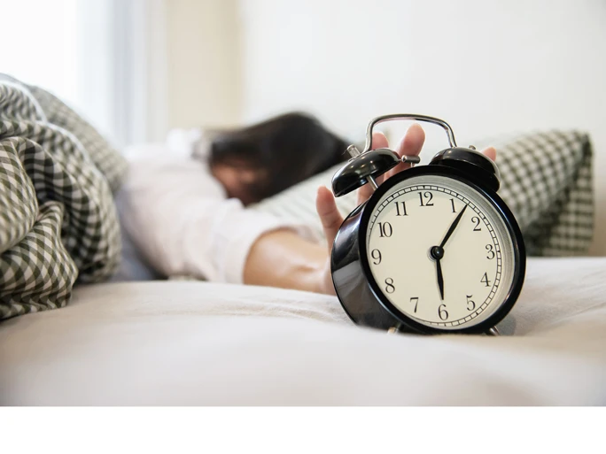 Symbolbild zum Beitrag Probleme nach der Zeitumstellung - Person im Bett mit einem Wecker.