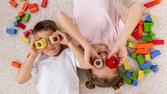 Zwei Kinder liegen am Boden und schauen durch Bauklötze. Daneben liegen noch weitere Klötze.