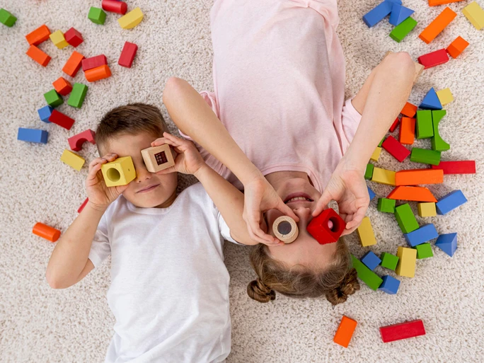 Zwei Kinder liegen am Boden und schauen durch Bauklötze. Daneben liegen noch weitere Klötze.