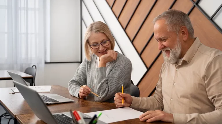 Eine Frau und ein Mann mittleren Alters sitzen zusammen vor ihren Laptops und fassen Ideen auf einem Papier zusammen
