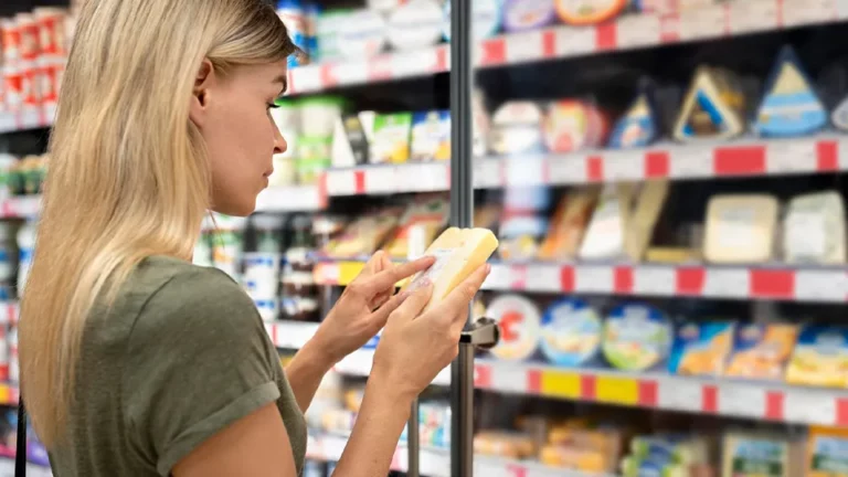 Frau steht an Kühltheke und liest die Nährstoffangabe auf einem Käse-Stück