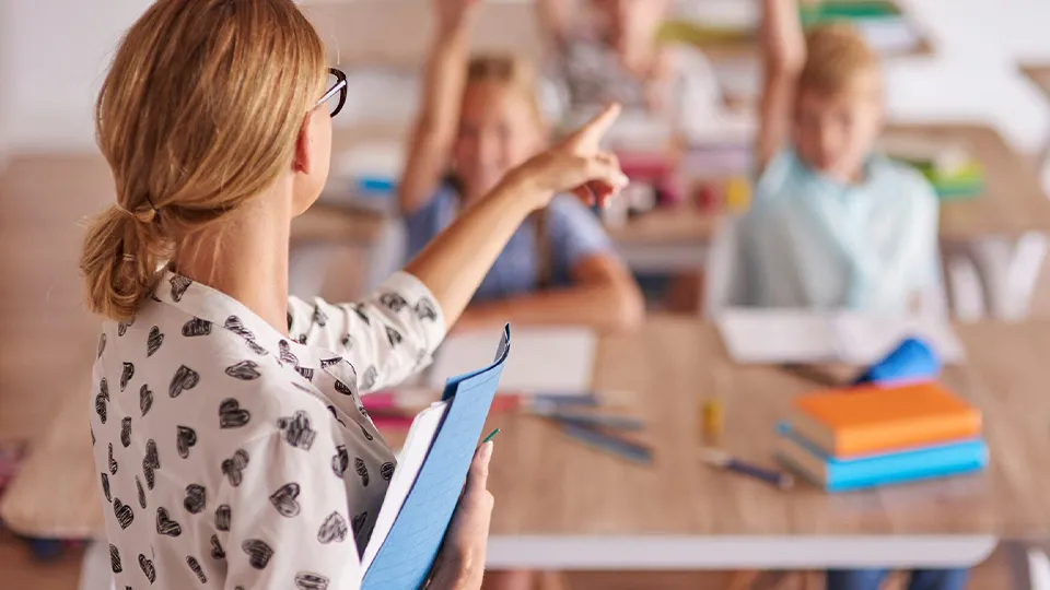 Lehrerin zeigt mit Finger auf die Schüler, die sich im Hintergrund melden.