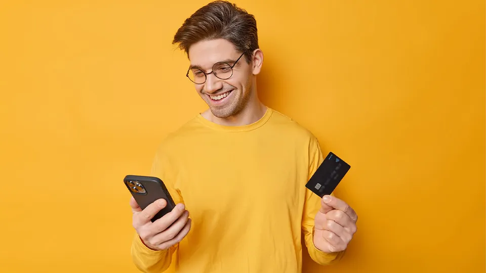 Junger Mann in einem gelben T-SHirt vor einer gelben Wand. Er hält eine Kreditkarte und ein Handy in den Händen.