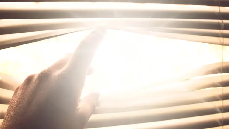 Hand öffnet mit den Fingern die Lamellen eines Sonnenschutzes. Die Sonne scheint grell in das Fenster.