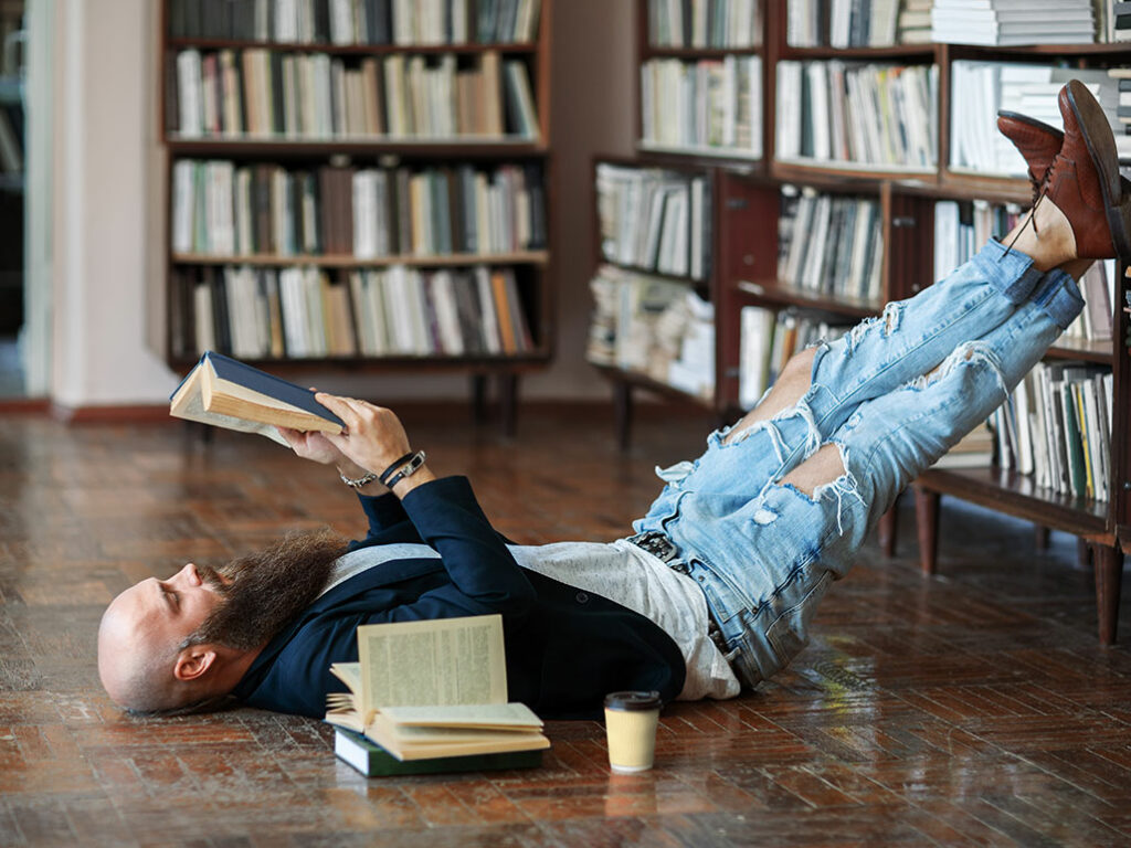 Mann liegend und lesend in der Bibliothek mit Füßen im Buchregal