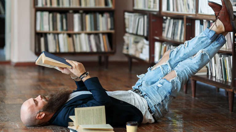 Mann liegend und lesend in der Bibliothek mit Füßen im Buchregal