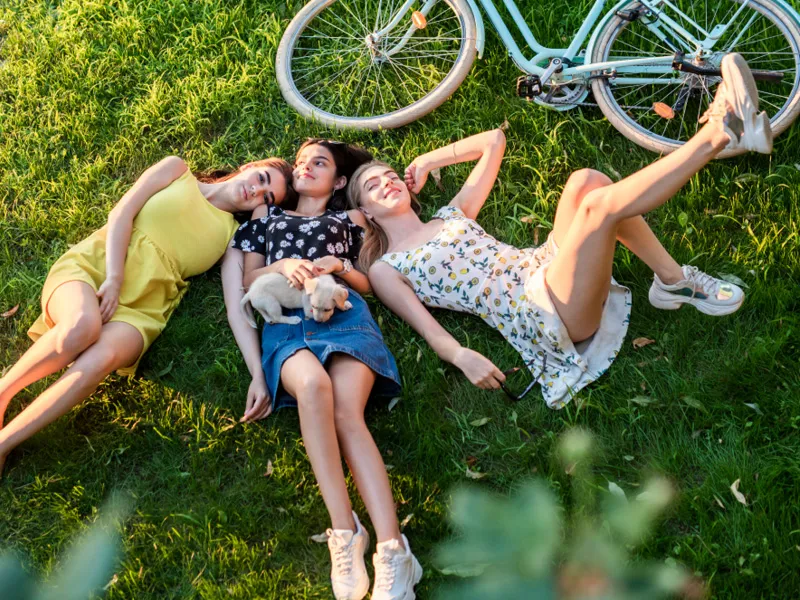 Drei Frauen liegen auf einer sommerlichen grünen Wiese neben einem Fahrrad.