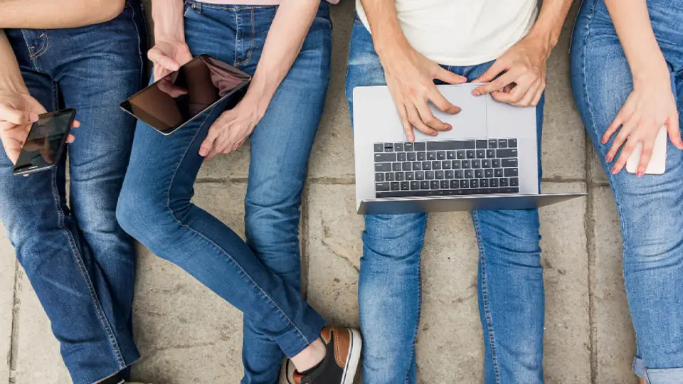 Vier Personen in Jeans sitzen am Boden un nutzen diverse Geräte wie Handy, Tablet und Laptop.