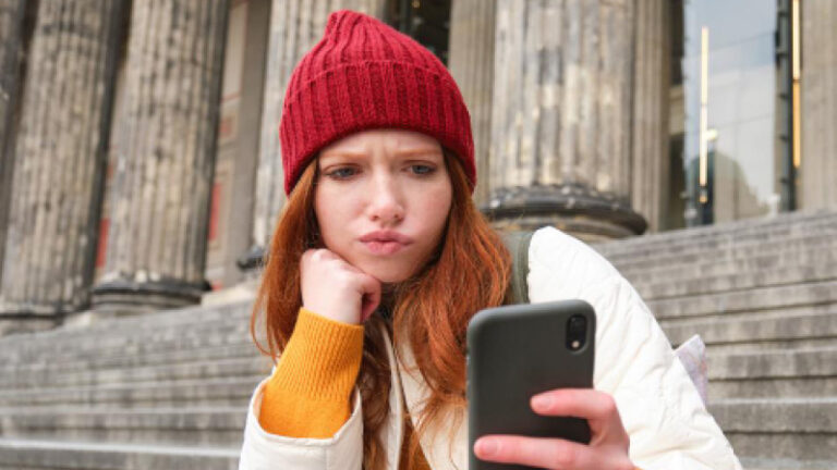 Junge Frau mit roter Mütze blickt kritisch auf ihr Smartphone.
