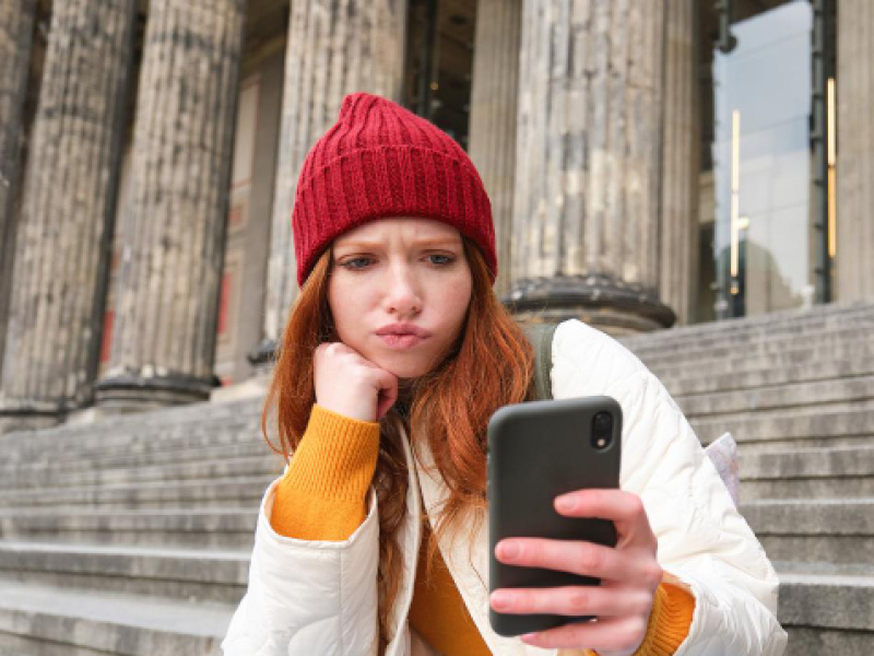 Junge Frau mit roter Mütze blickt kritisch auf ihr Smartphone.