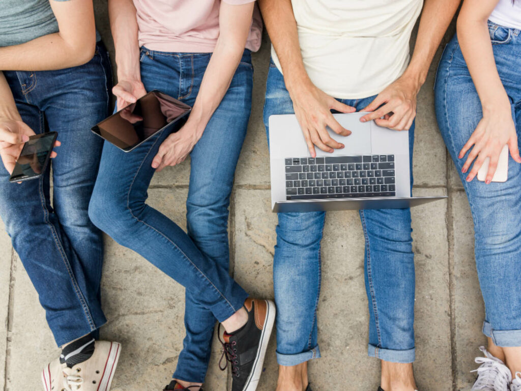 Vier Personen sitzen auf dem Boden und nutzen verschiedene Mobilgeräte und Laptops
