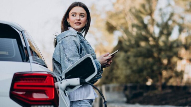 Eine Frau lehnt sich entspannt an ein Auto, das gerade an einer Ladestation angeschlossen ist.