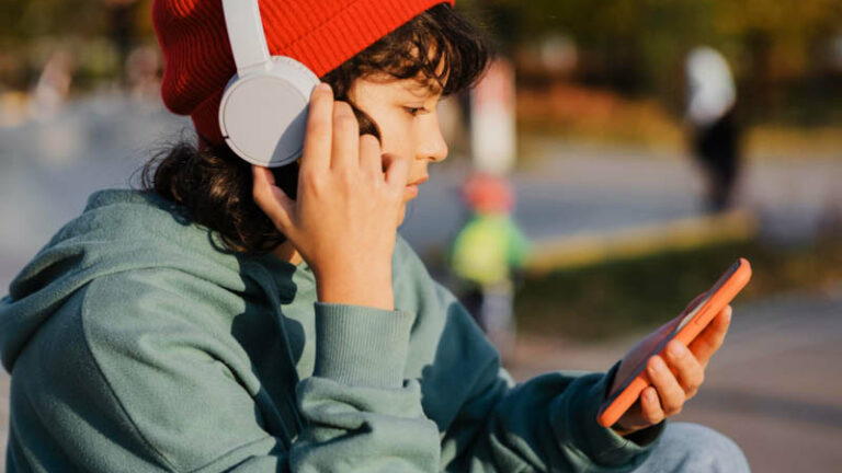 Ein eJugendliche mit roter Mütze und Over-Ear-Kopfhörern hört Musik und hält ein Mobiltelefon in der Hand.