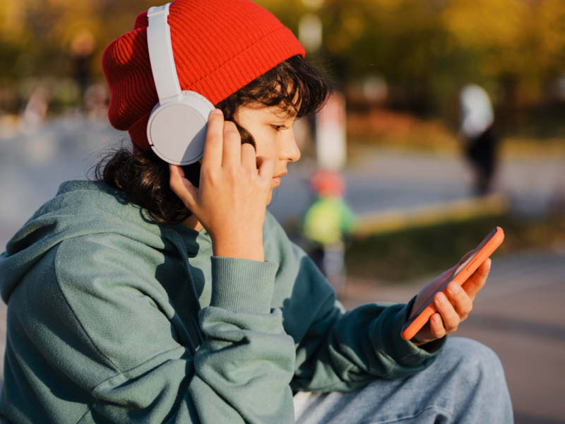 Ein eJugendliche mit roter Mütze und Over-Ear-Kopfhörern hört Musik und hält ein Mobiltelefon in der Hand.