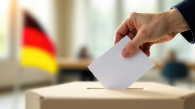 Hand schmeißt Wahlzettel in Wahlurne, Deutschlandflagge unscharf im Hintergrund