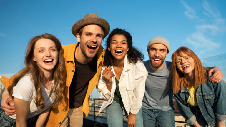 Fünf junge Menschen lachen fröhlich zusammen vor blauem Himmel.