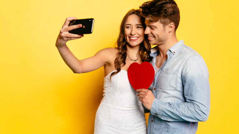 Junges Paar macht ein Selfie, sie trägt ein weißes Kleid, er hält ein rotes Papierherz in der Hand.