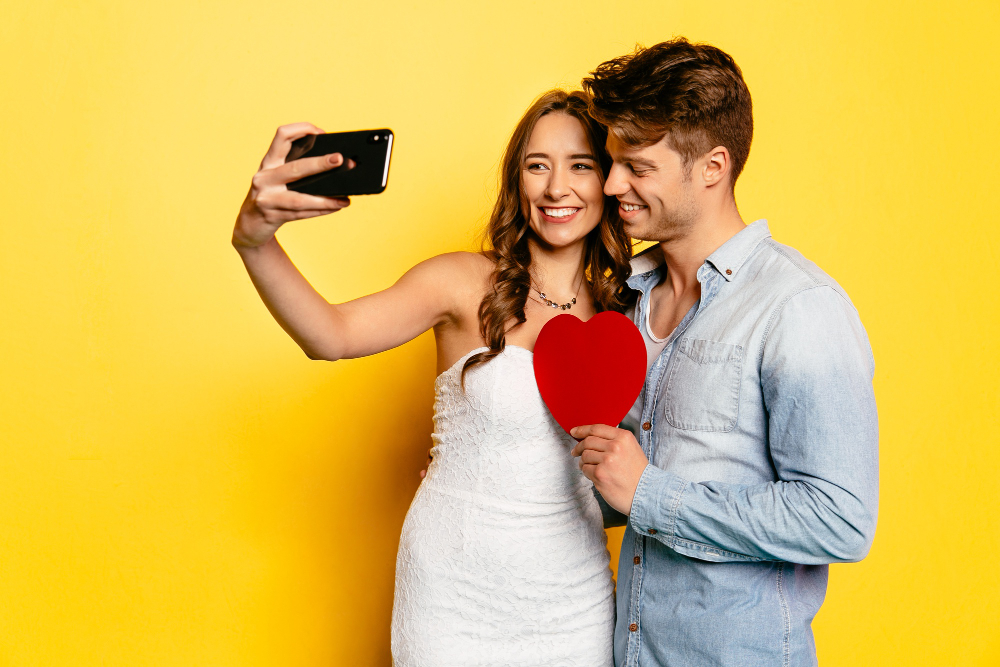 Junges Paar macht ein Selfie, sie trägt ein weißes Kleid, er hält ein rotes Papierherz in der Hand.