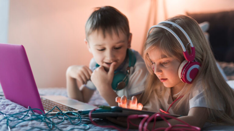 Zwei Kinder liegen im Bett, nutzen Laptop und Tablet, eins trägt Kopfhörer.