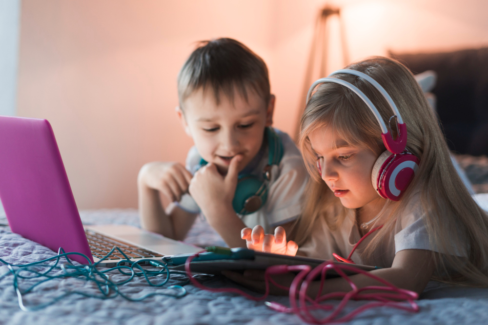 Zwei Kinder liegen im Bett, nutzen Laptop und Tablet, eins trägt Kopfhörer.