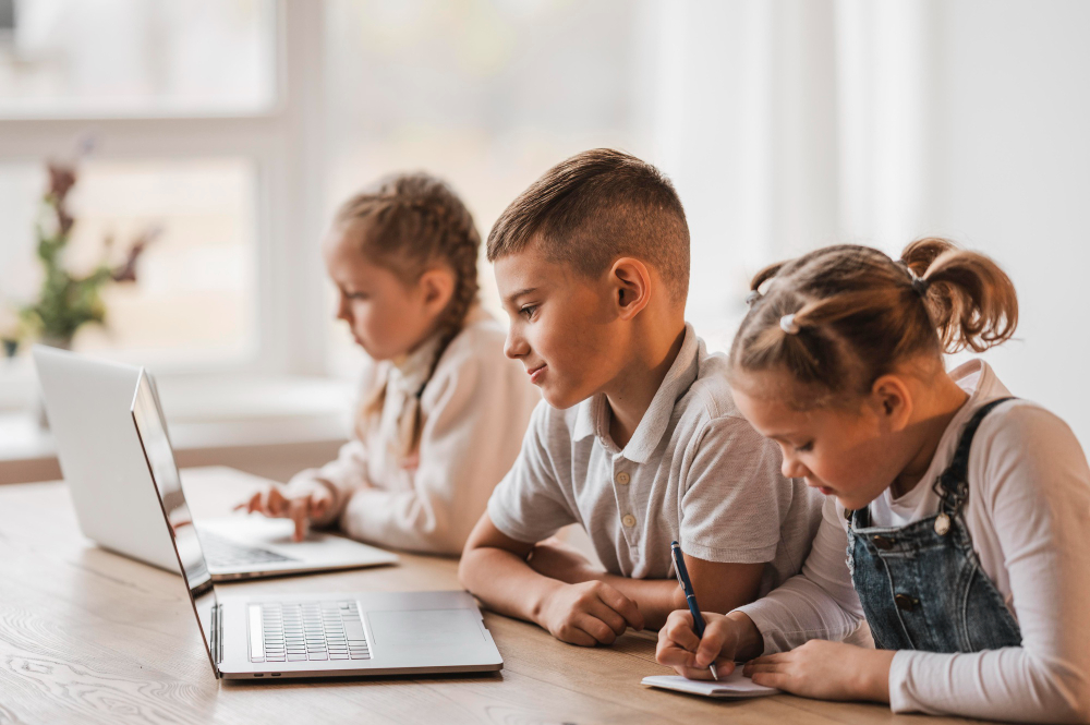 Drei Kinder arbeiten an Laptops, eins schreibt auf einem Notizblock.