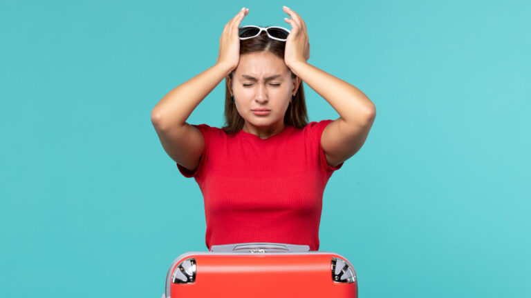 Gestresste Frau hält sich den Kopf, Koffer steht neben ihr – Symbolbild für Stress im Urlaub